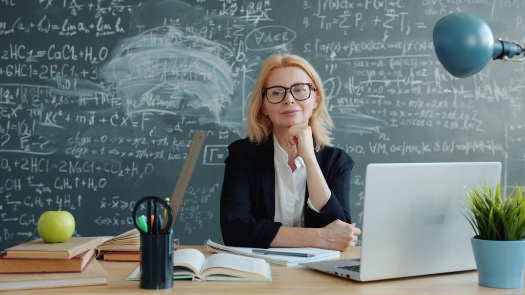 Woman in glasses at desk with laptop and chalkboard.