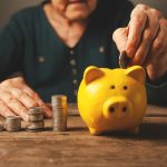 Elderly hands depositing coins into a yellow piggy bank.