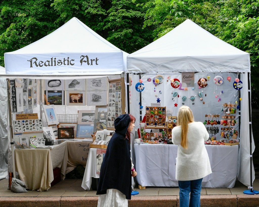 Two women browse art and crafts at an outdoor market.