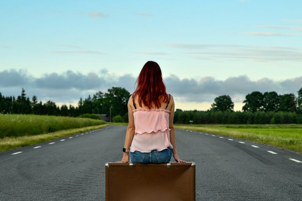 Red-haired woman sits on suitcase on empty road.