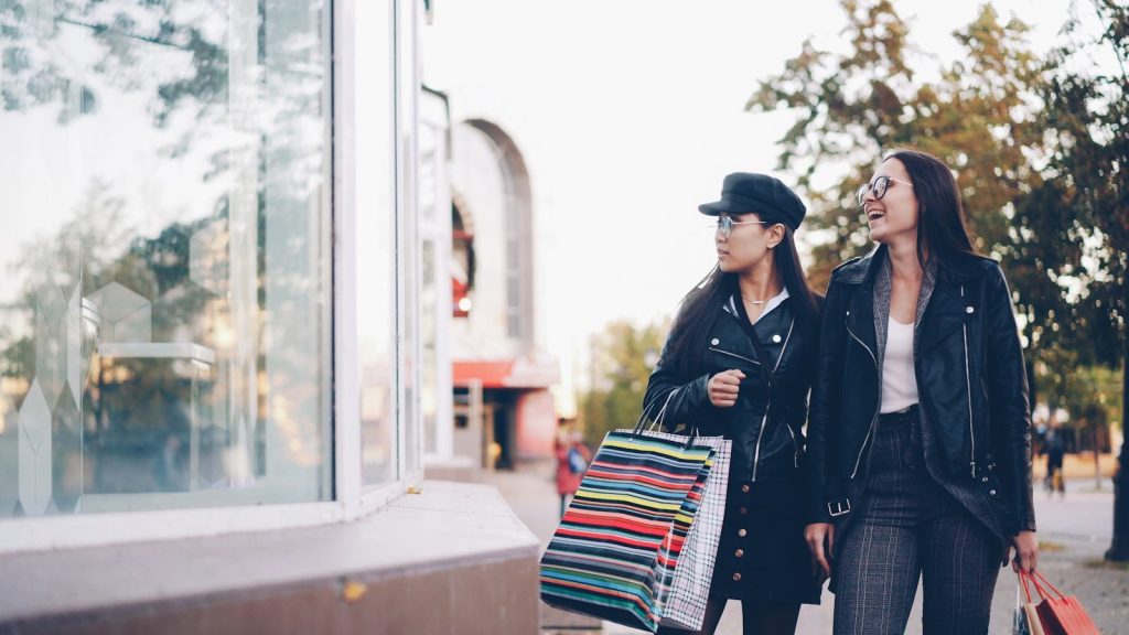 Two women walking with shopping bags past a storefront.