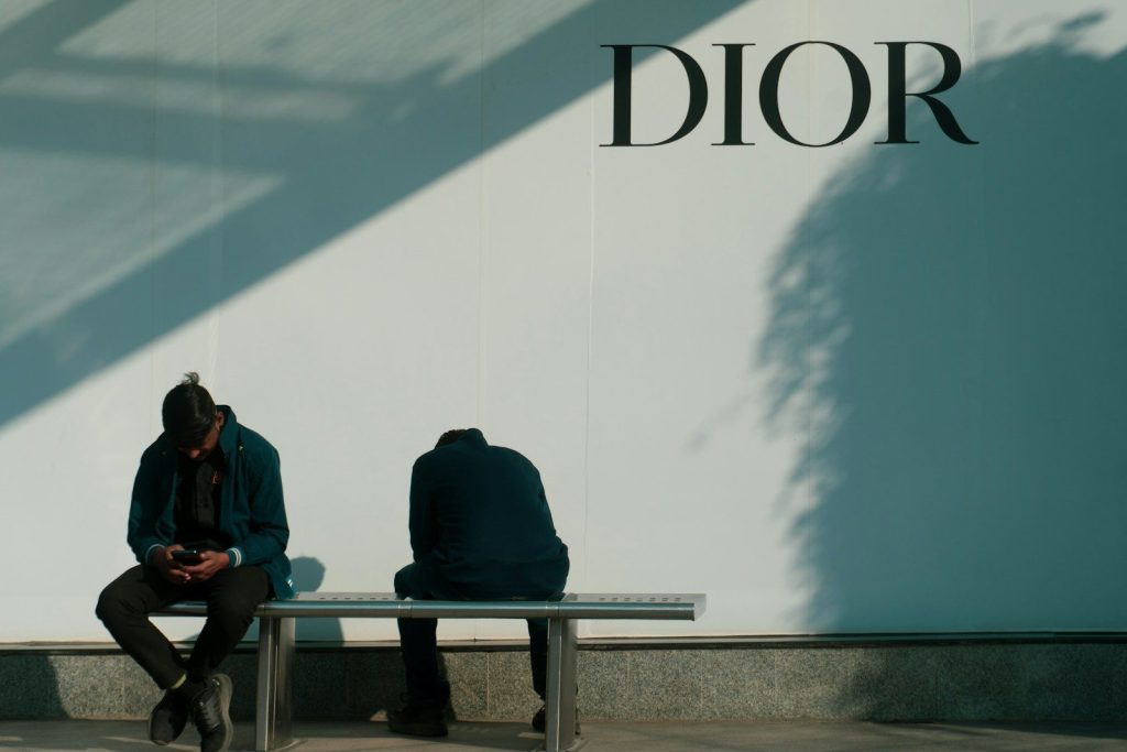 Two people sit on a bench near a dior sign.