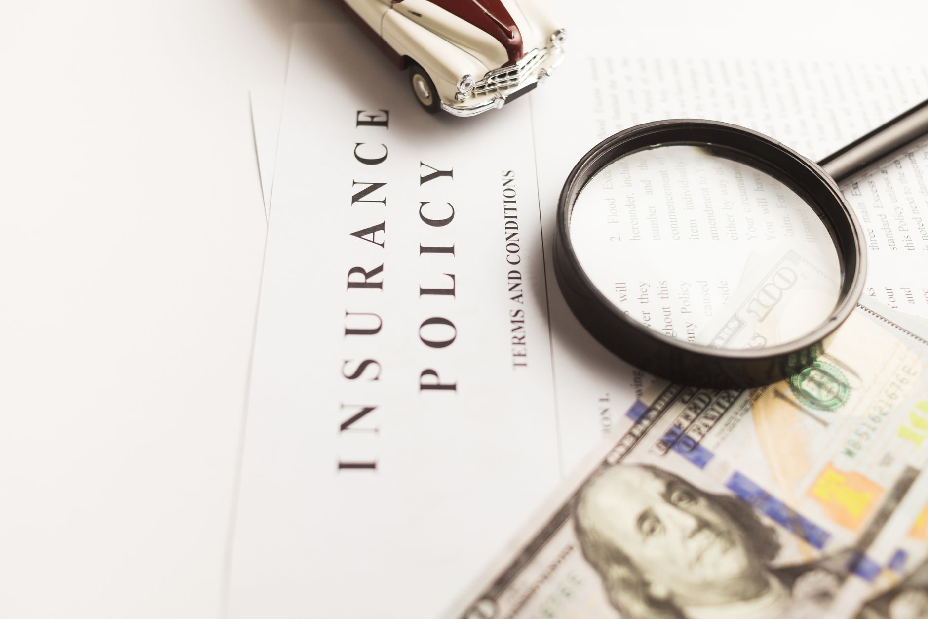 Close up image of an insurance policy with a magnifying glass, money, and toy car.