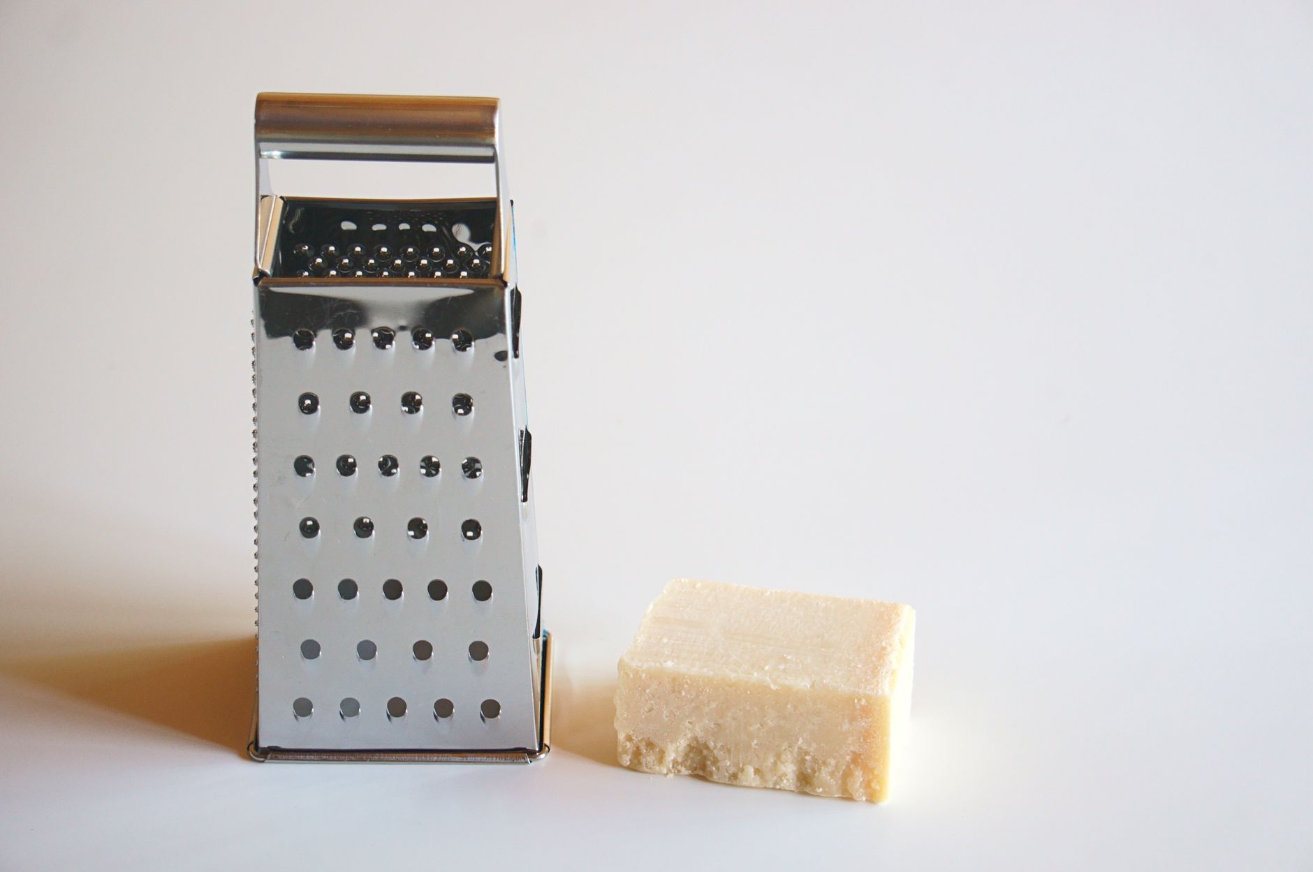 A stainless steel grater next to a block of parmesan cheese on a white surface.