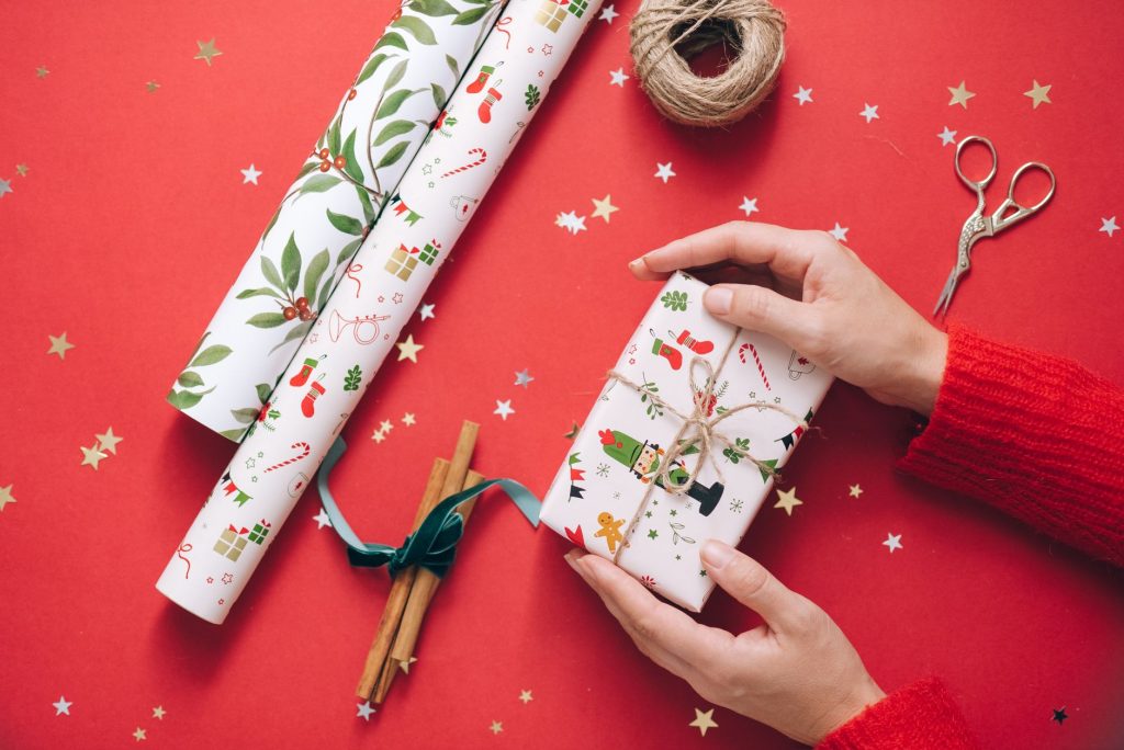 Hands wrapping a Christmas present on a vibrant red surface with festive decorations.