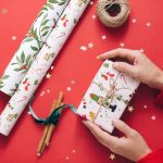 Hands wrapping a Christmas present on a vibrant red surface with festive decorations.
