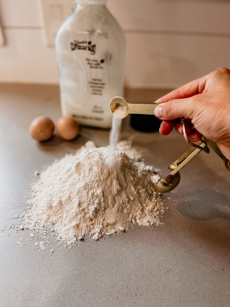 Hand pouring salt onto flour for baking with eggs in the background.