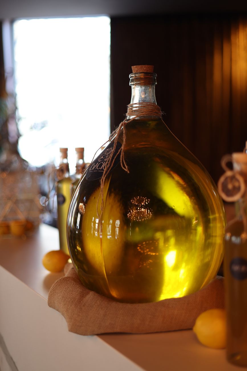 A large glass bottle filled with olive oil on a rustic bar counter with lemons beside it.