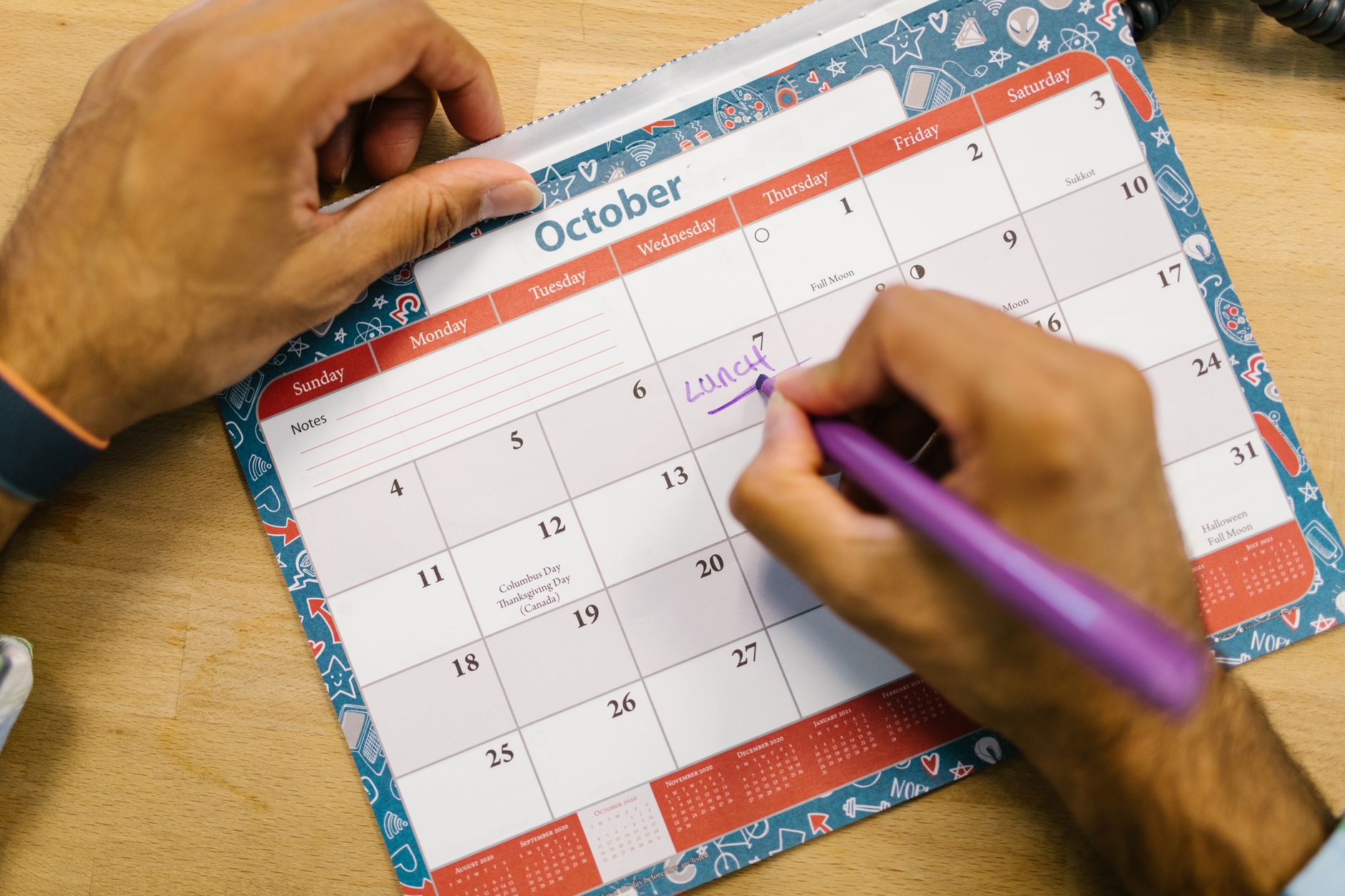 Close up of a person writing a lunch reminder on an October calendar with a purple pen.