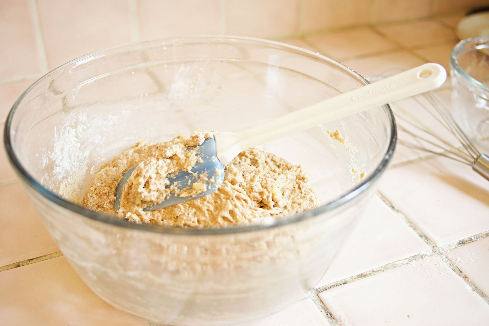 A close up image of dough being mixed in a glass bowl with a white spatula on a tiled surface.