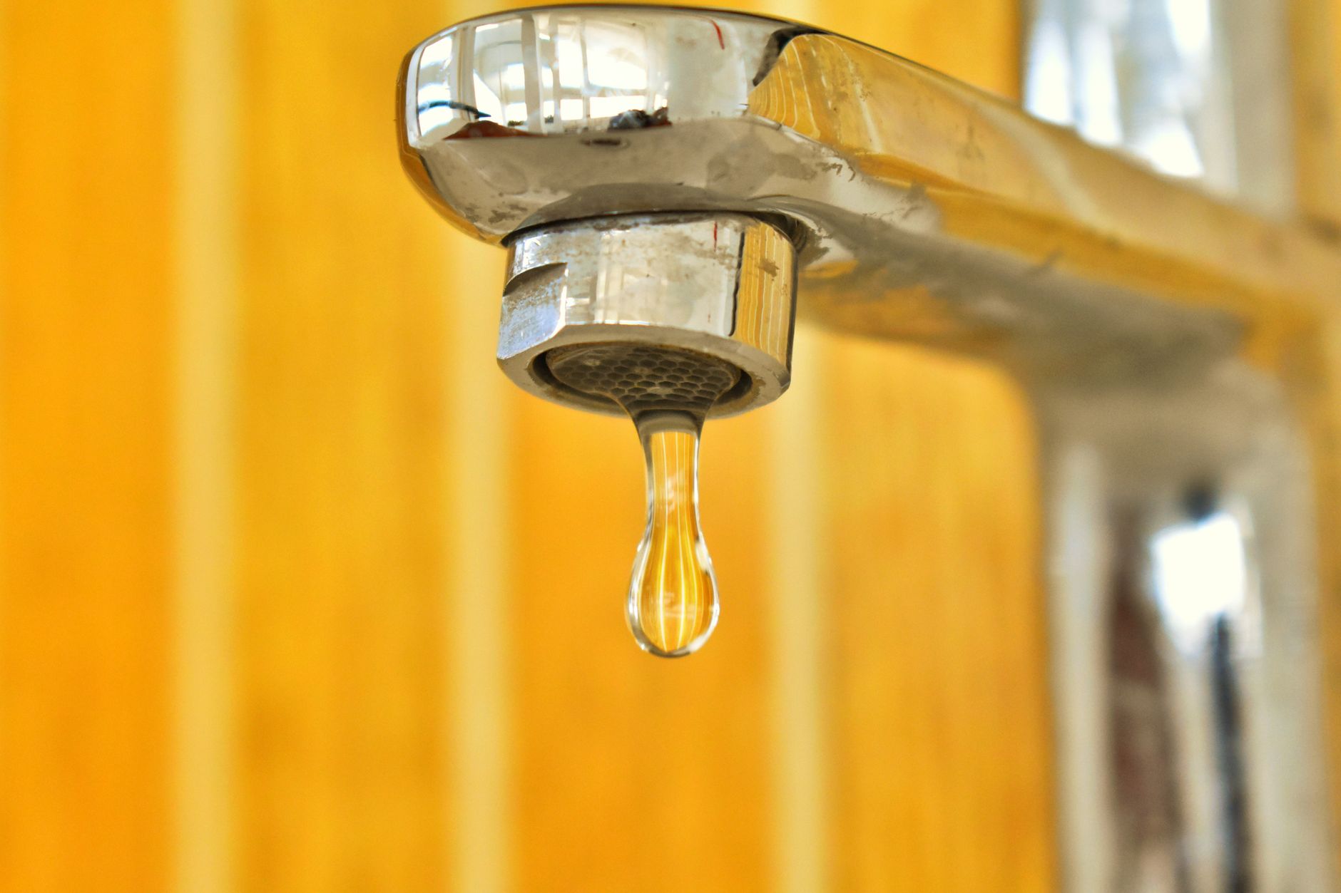 A close up shot of a water droplet hanging from a stainless steel faucet, highlighting surface tension.