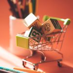 A miniature shopping cart holds alphabet blocks beside colored pencils on a warm background.