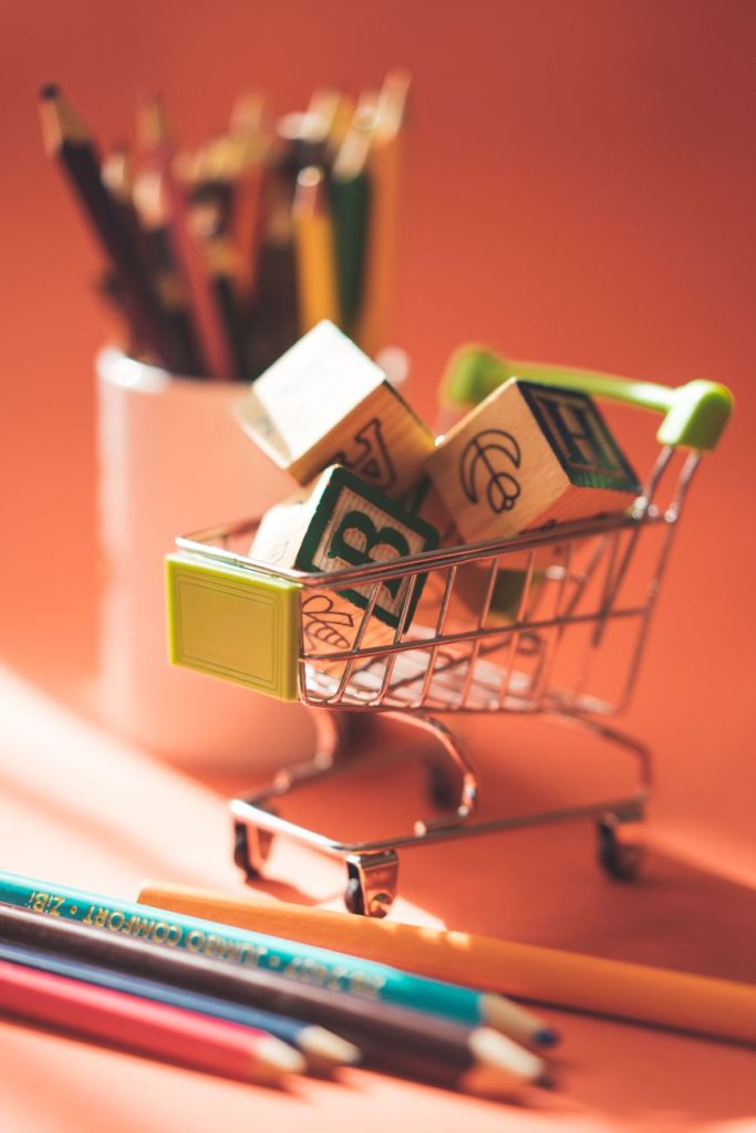 A miniature shopping cart holds alphabet blocks beside colored pencils on a warm background.