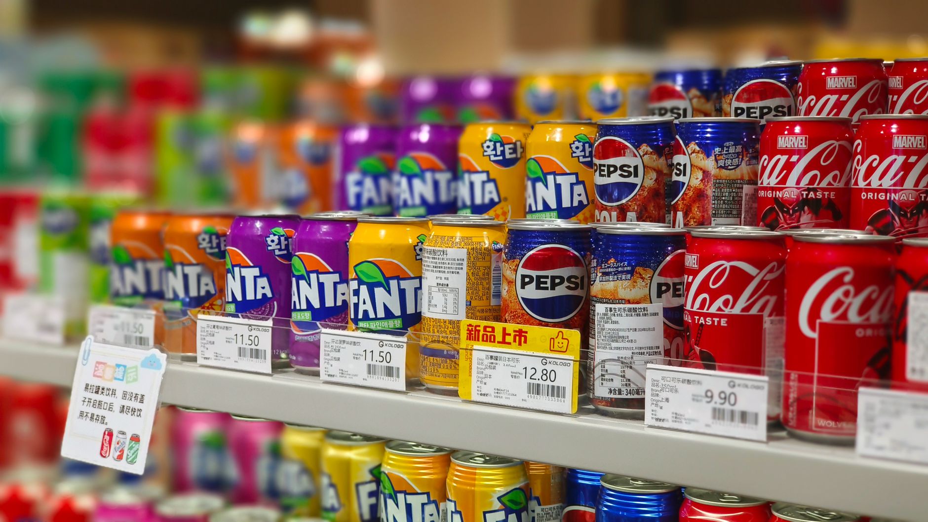 Colorful soda cans including Fanta, Pepsi, and Coca Cola arranged on supermarket shelves, price tags visible.