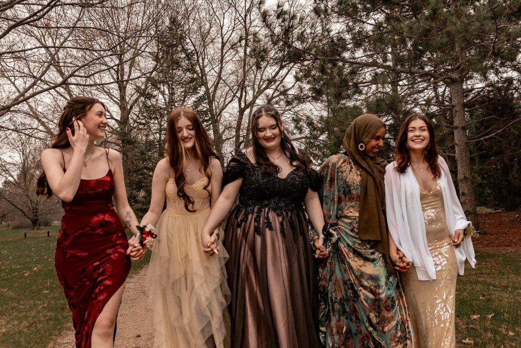 Five smiling women in formal dresses holding hands outdoors.