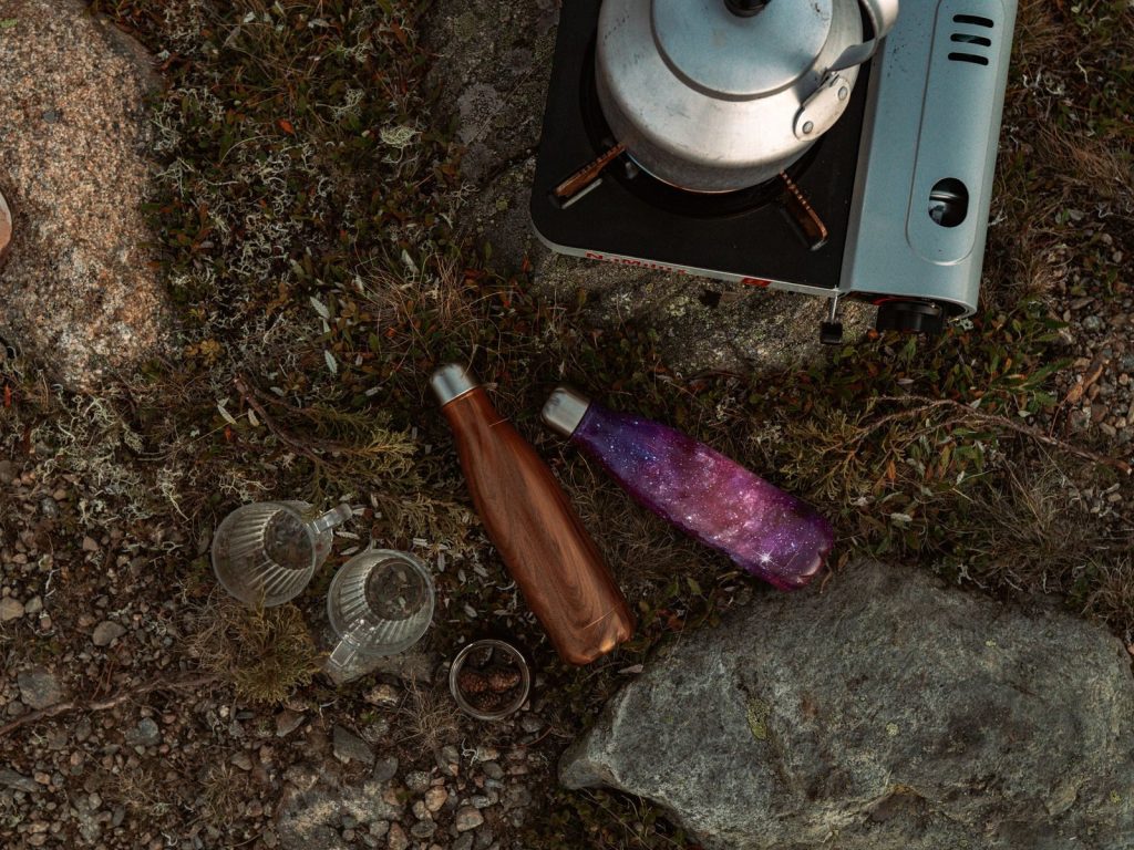 Top view of camping essentials on grassy ground, including a kettle and bottles.