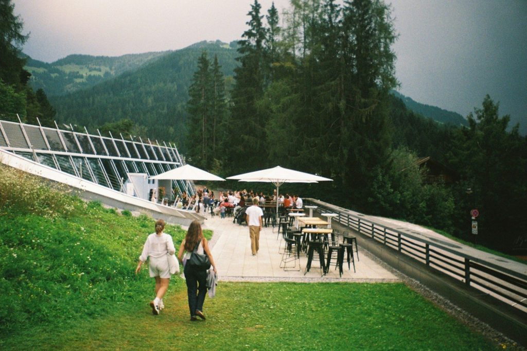 People walk past an outdoor cafe with mountains in background.