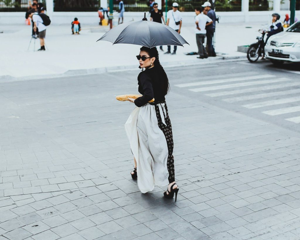 woman wearing black long-sleeved shirt and sunglasses using umbrella while walking on pathway and other people standing near road during daytime