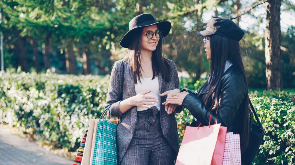 Two stylish women with shopping bags talking outdoors.