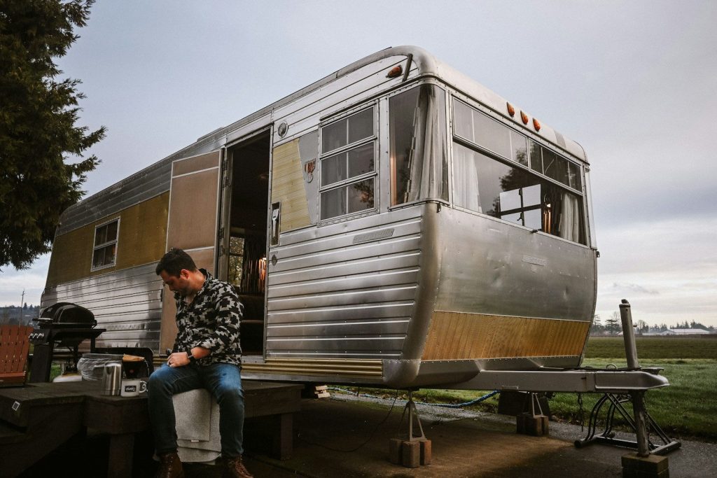 Man sits by a vintage silver and gold travel trailer.