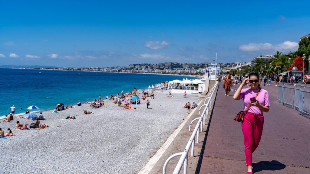 Woman walks on promenade beside crowded beach and blue sea.