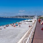 Woman walks on promenade beside crowded beach and blue sea.